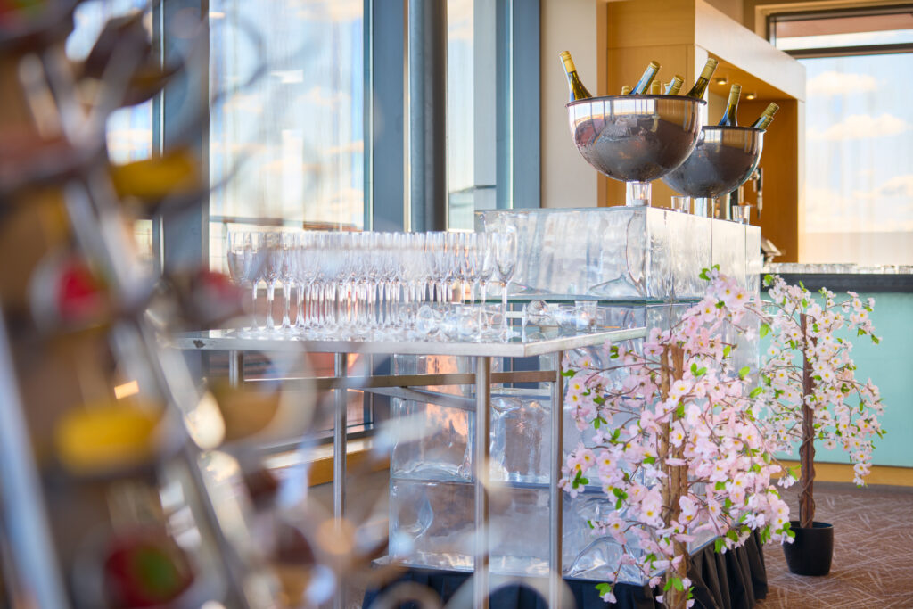 A table with empty champagne glasses, two ice buckets with champagne bottles, and pink artificial cherry blossom decorations in a bright room with large windows.