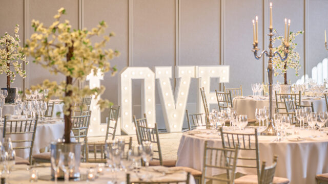 Elegant banquet hall with round tables, gold chairs, floral centerpieces, and a large illuminated