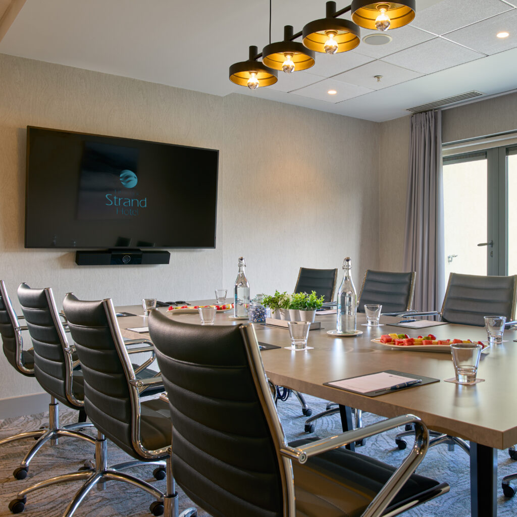 Modern conference room with a long table, black leather chairs, bottled water, notepads, and a large wall-mounted screen displaying 