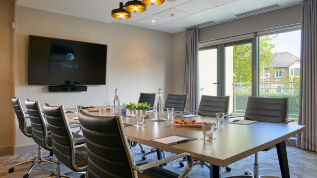 Modern conference room with a long table, black chairs, water bottles, notepads, and fruit plates, featuring a TV screen and large window with curtains overlooking greenery.