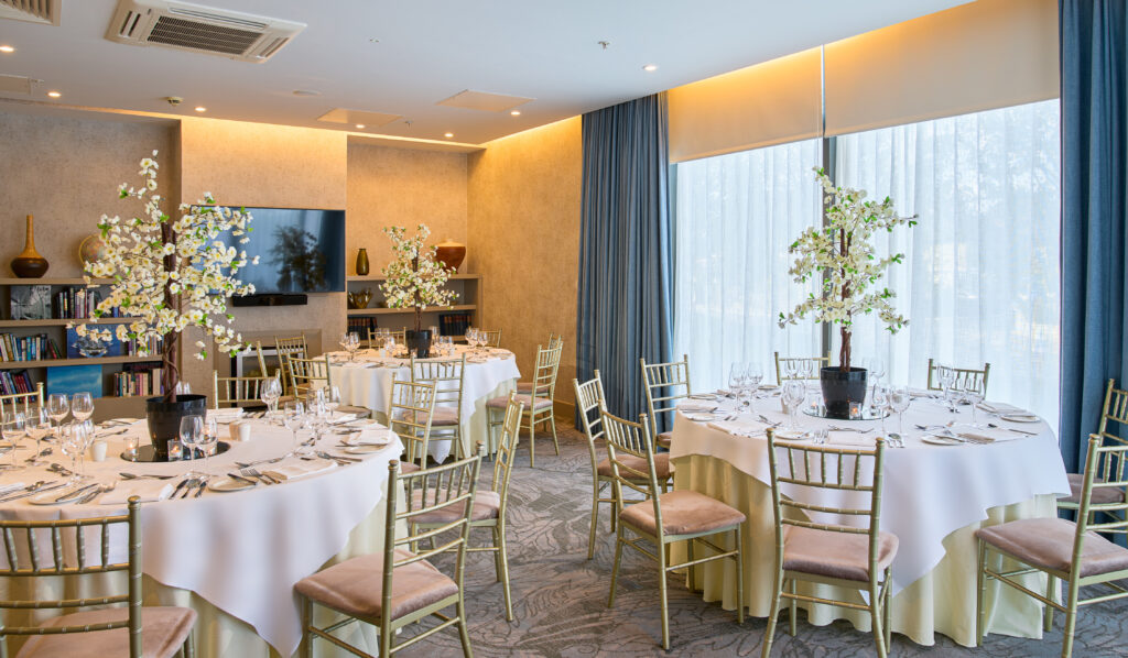Formal dining room with round tables covered in white cloths, gold chairs, elegant table settings, and decorative trees as centerpieces. Large windows let in natural light.