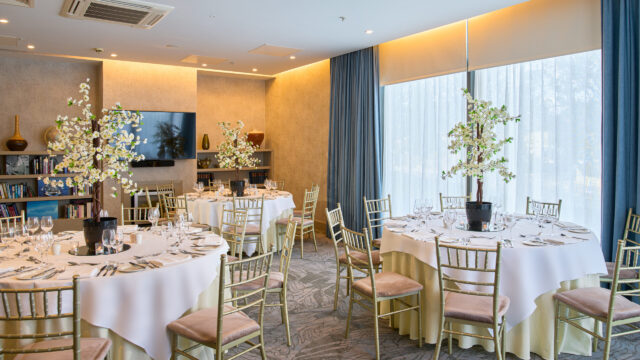 Formal dining room with round tables covered in white cloths, gold chairs, elegant table settings, and decorative trees as centerpieces. Large windows let in natural light.