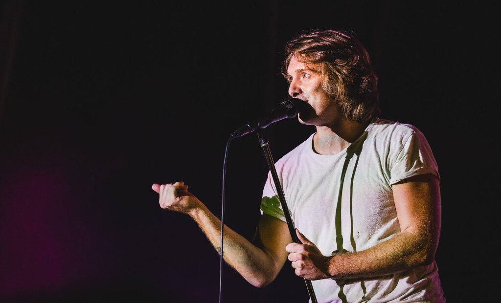 A person in a white t-shirt stands on stage, speaking or singing into a microphone, with one hand gesturing to the side.