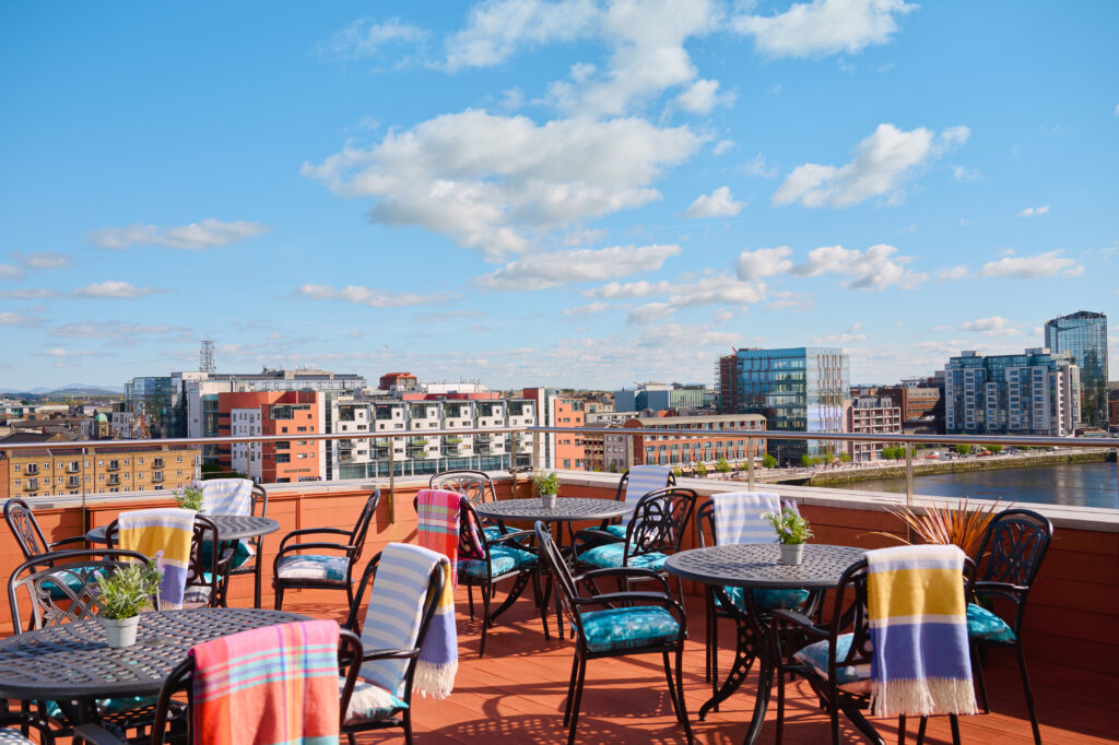 Rooftop terrace with tables and chairs, some with colorful blankets, overlooking a cityscape and river under a blue sky with scattered clouds.