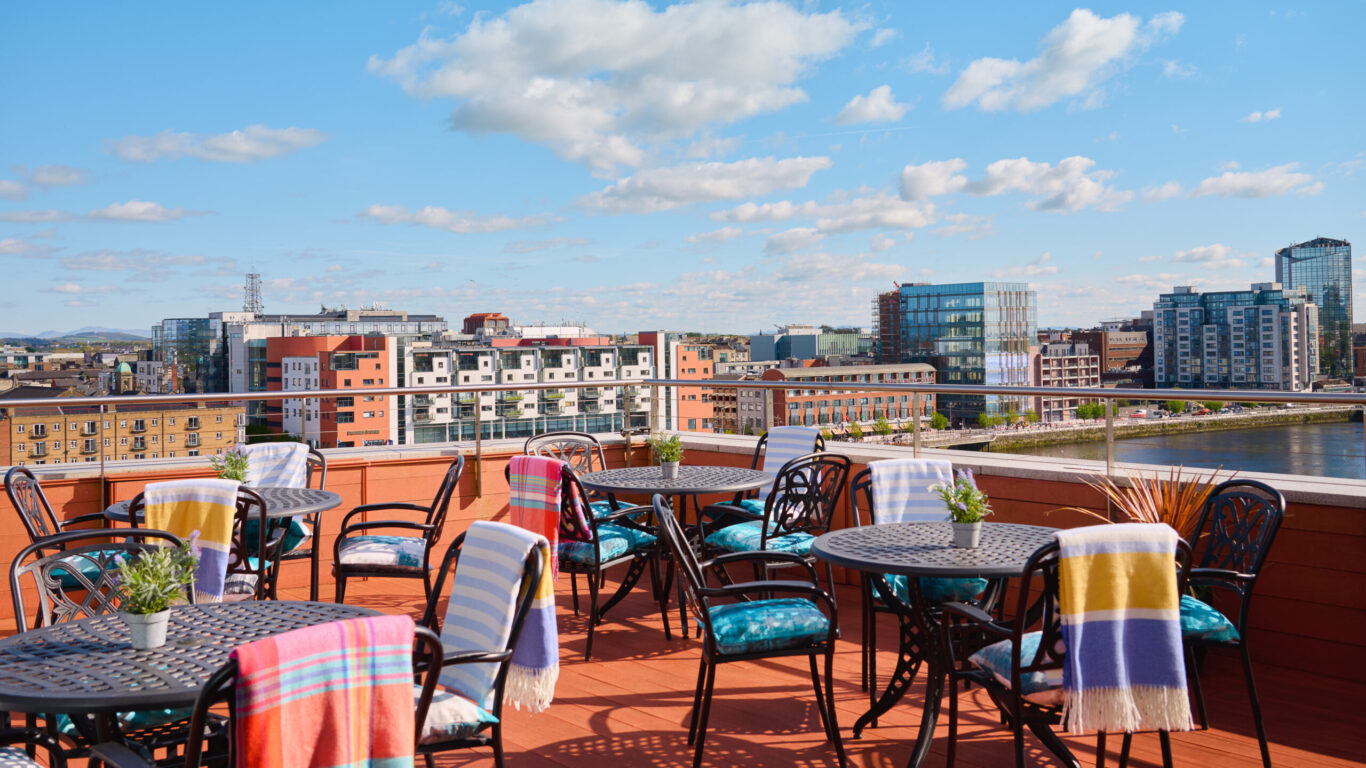 Rooftop terrace with tables and chairs, some with colorful blankets, overlooking a cityscape and river under a blue sky with scattered clouds.