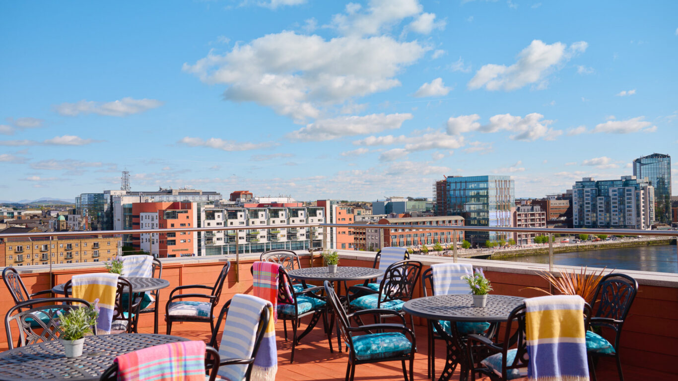 Rooftop terrace with tables and chairs, some with colorful blankets, overlooking a cityscape and river under a blue sky with scattered clouds.