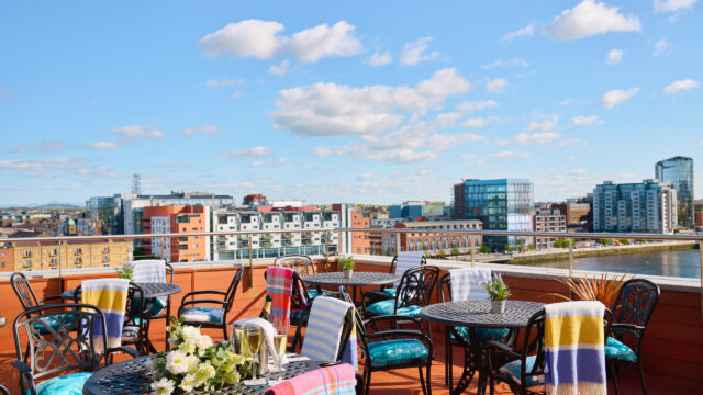 Outdoor rooftop patio with metal tables and chairs, colorful blankets, and a cityscape view under a blue sky with scattered clouds.