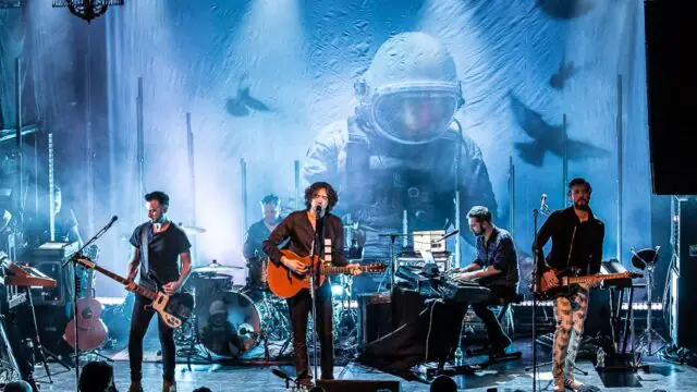 A band performs on stage with guitars, drums, and keyboards; a large backdrop features an astronaut illustration under blue stage lighting.