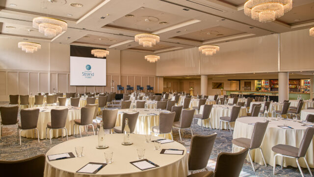 Spacious banquet hall set up for a conference with round tables, chairs, notepads, water bottles, a stage, projection screen, and chandeliers.