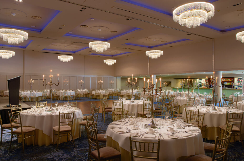 A large banquet hall set for an event with round tables, formal place settings, candelabra centerpieces, chandeliers, and a bar in the background.