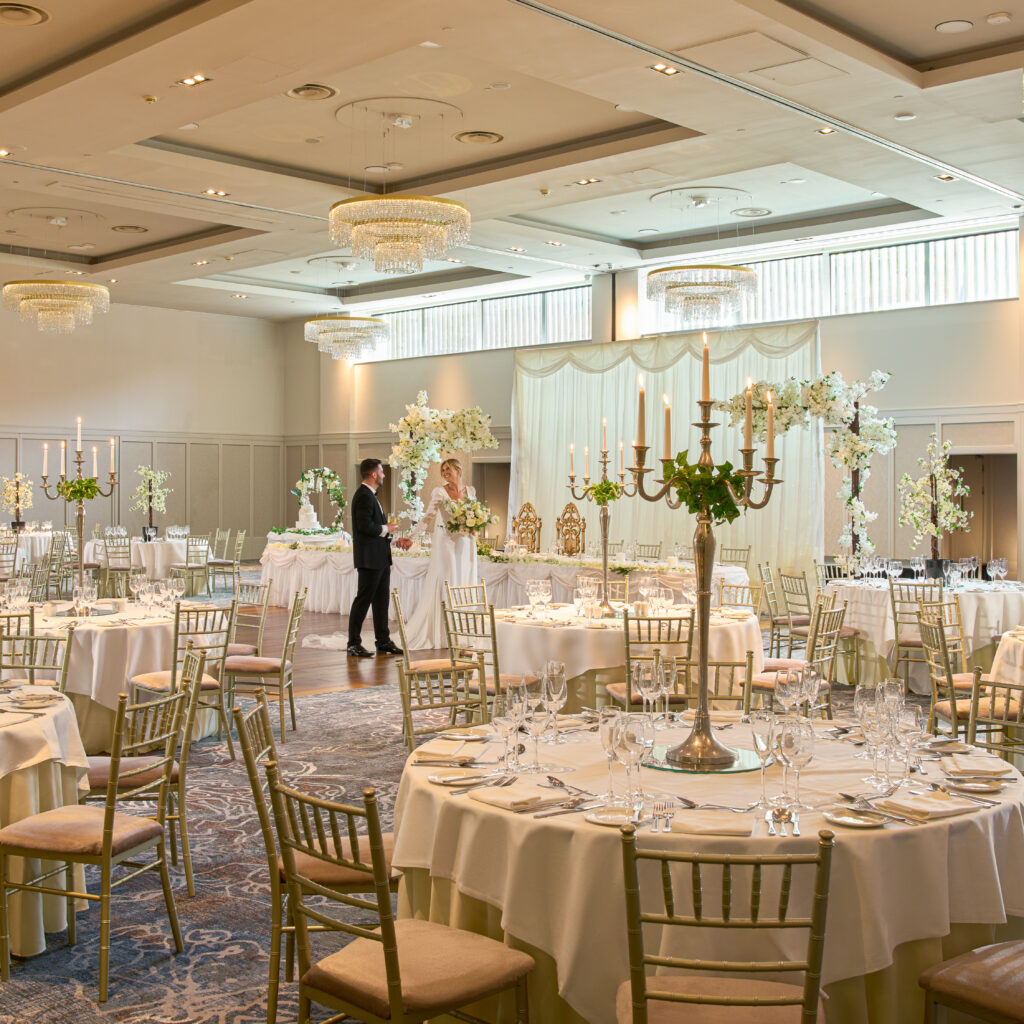 Elegant banquet hall set for a wedding reception with round tables, gold chairs, floral centerpieces, and two people standing near a decorated stage.