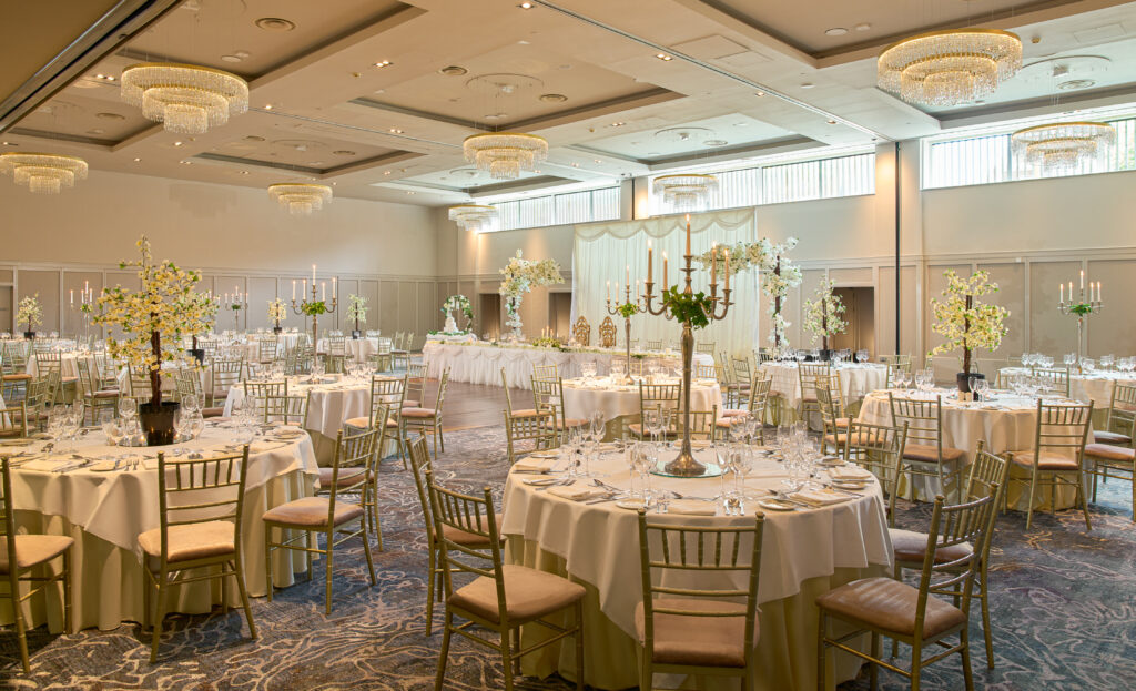 Elegant banquet hall set up for an event with round tables, white tablecloths, gold chairs, floral centerpieces, and chandeliers overhead.