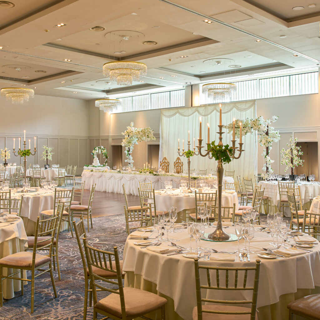 Elegant banquet hall set for an event with round tables, gold chairs, white tablecloths, candelabras, and floral centerpieces; a decorated head table is at the front.