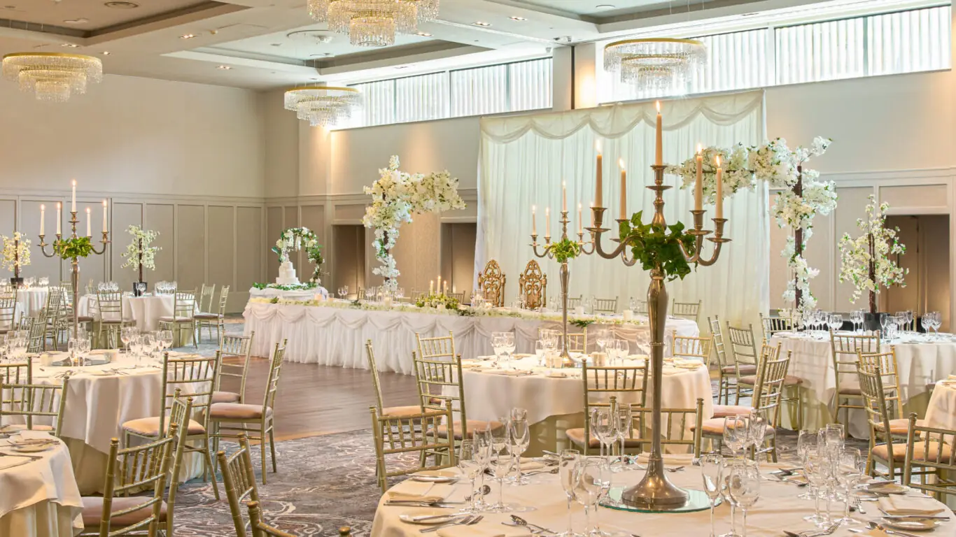 Elegant banquet hall set for an event with round tables, gold chairs, white tablecloths, candelabras, and floral centerpieces; a decorated head table is at the front.