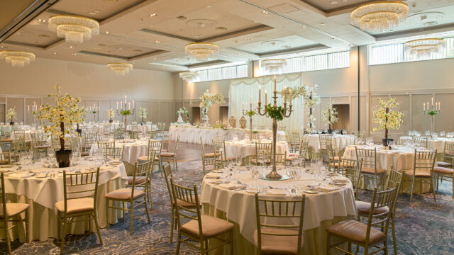 Elegant banquet hall set up for an event with round tables, white tablecloths, gold chairs, floral centerpieces, and chandeliers overhead.