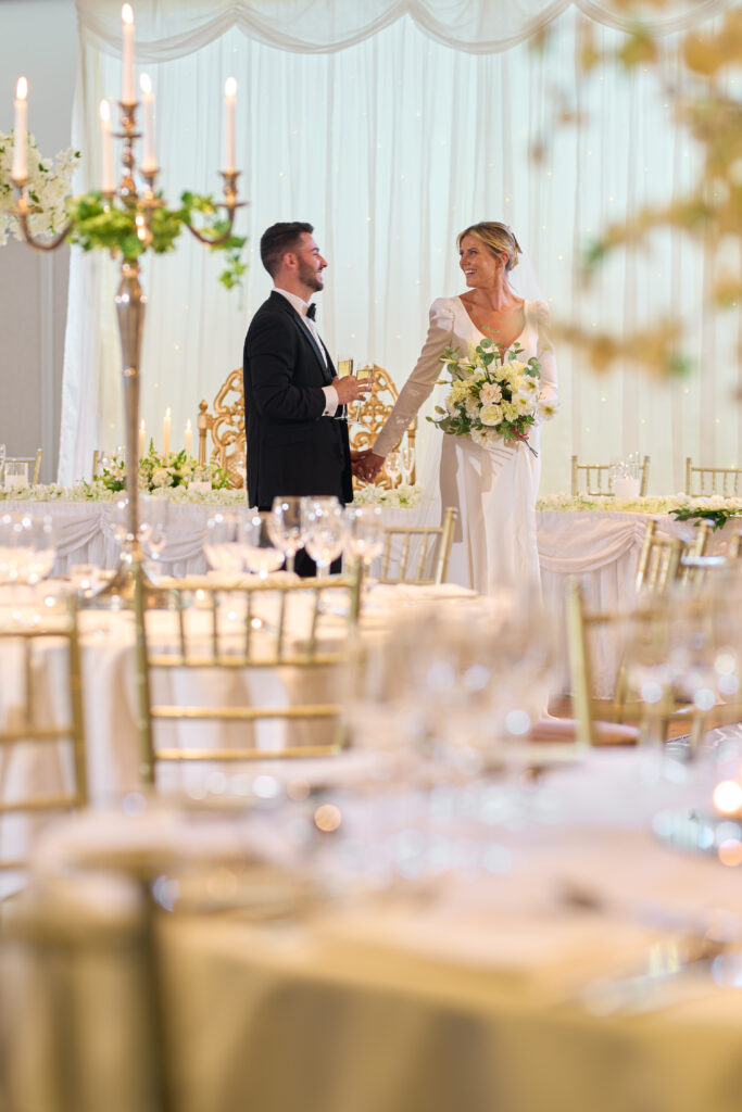 A bride and groom stand holding hands and smiling at each other in an elegantly decorated wedding reception hall with gold chairs and floral arrangements.