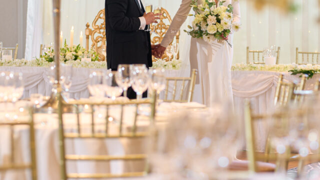 A bride and groom stand holding hands and smiling at each other in an elegantly decorated wedding reception hall with gold chairs and floral arrangements.