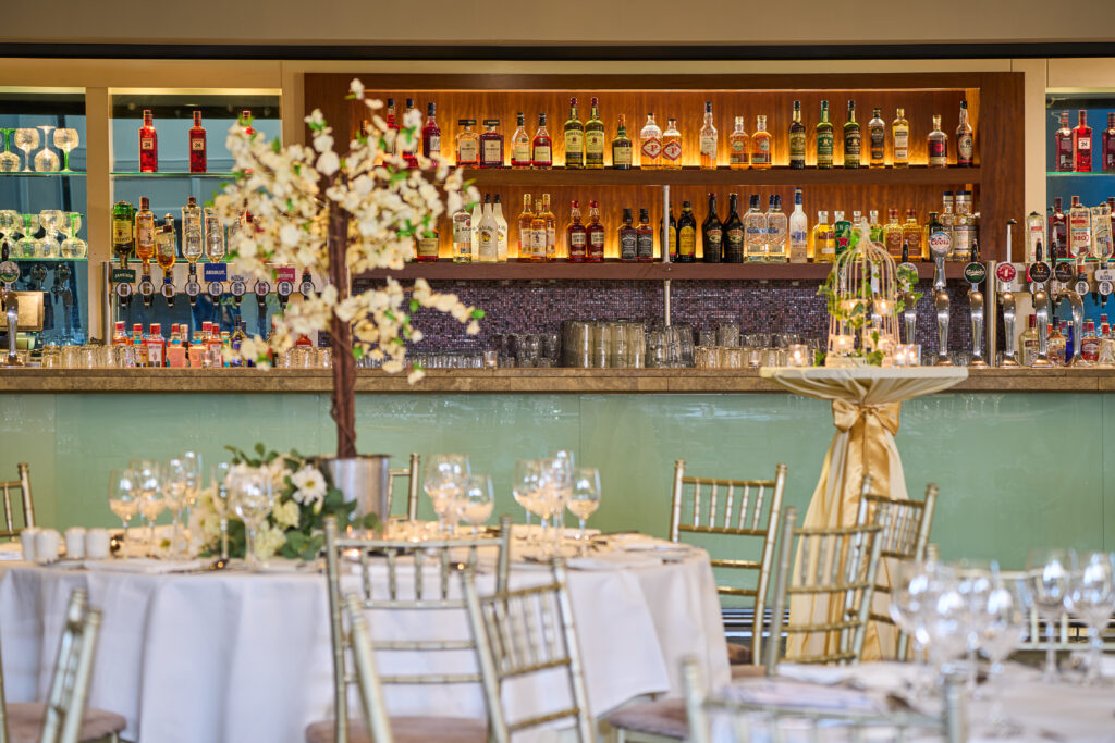 Elegant bar setup with a variety of spirits and glassware behind a counter; in the foreground, decorated round tables and gold chairs set for an event or celebration.