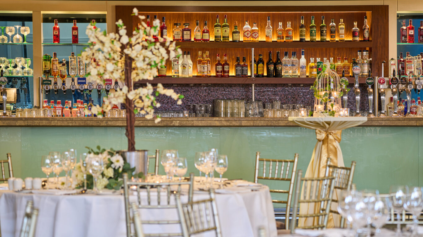 Elegant bar setup with a variety of spirits and glassware behind a counter; in the foreground, decorated round tables and gold chairs set for an event or celebration.