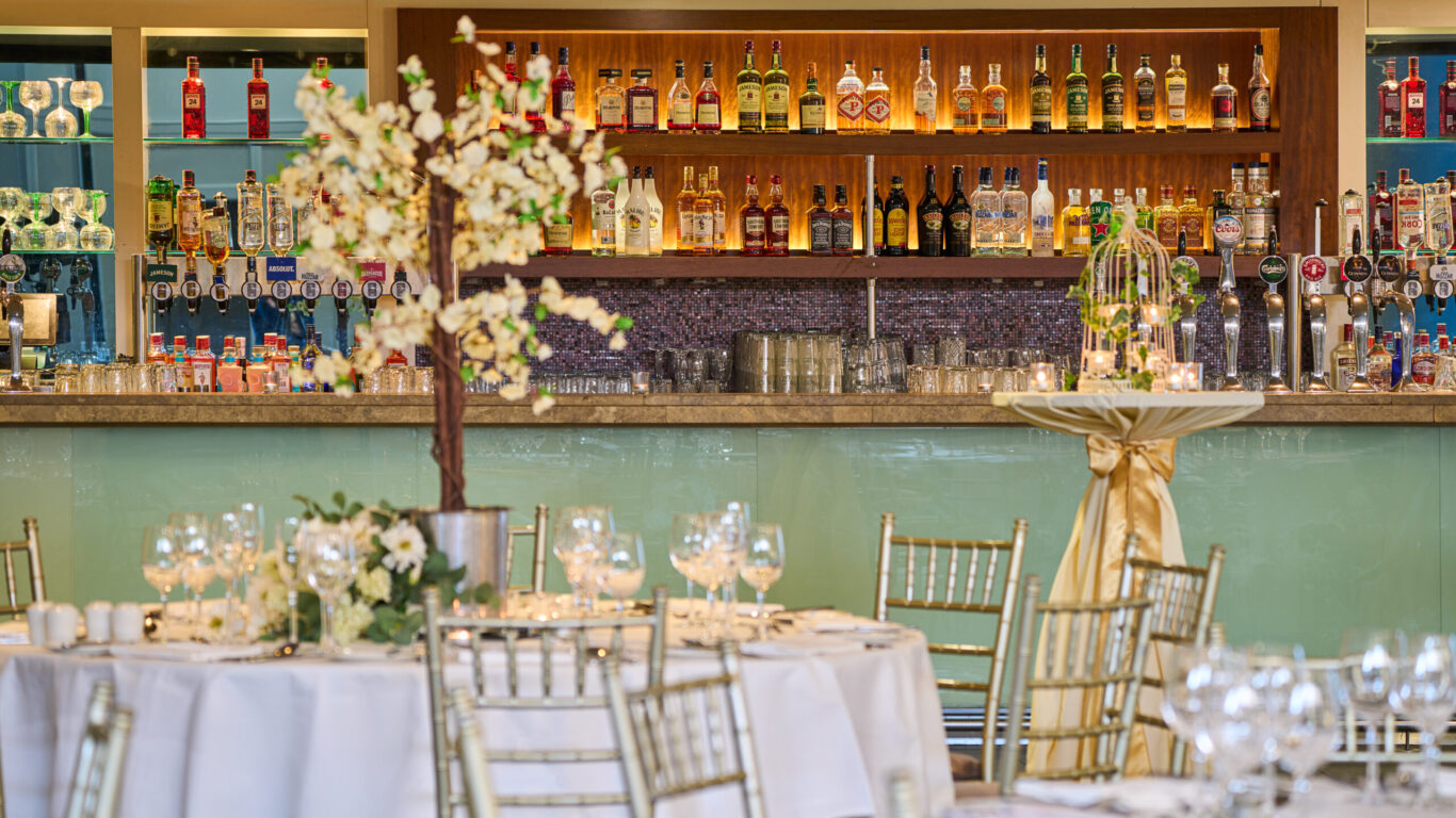 Elegant bar setup with a variety of spirits and glassware behind a counter; in the foreground, decorated round tables and gold chairs set for an event or celebration.