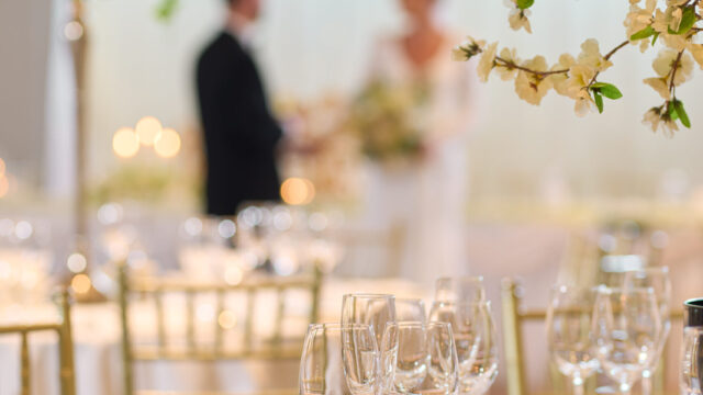 A decorated wedding reception table with glasses, plates, and cutlery in focus; a bride and groom stand blurred in the background.