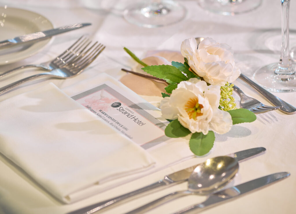 A neatly set dining table with silverware, a white napkin, a printed menu from Spark Hotel, and a small artificial white flower arrangement.