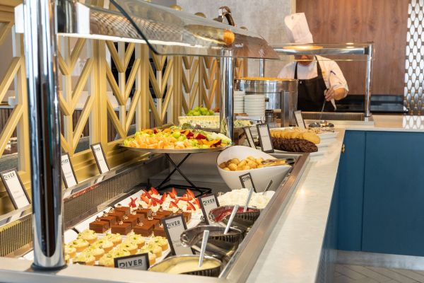 A buffet counter with various desserts, fruits, and toppings is displayed in front, while a chef in uniform works behind the counter.
