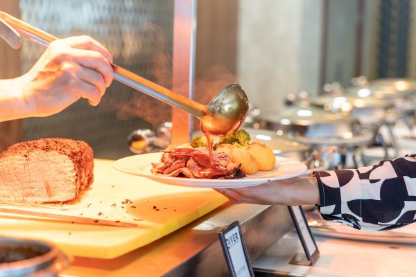 A person serves gravy onto a plate of sliced roast beef, vegetables, and potatoes at a buffet-style food station.