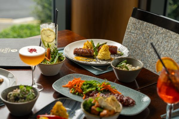 A restaurant table set with plated main dishes, sides, salads, and assorted cocktails near a window with natural light.