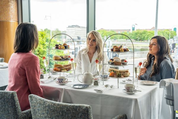 Three women sit at a table with tiered trays of afternoon tea food, teapots, and cups, next to a large window overlooking a cityscape.