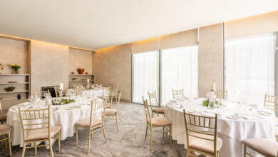 A bright banquet room with round tables set for dining, gold chairs, large windows with sheer curtains, and minimalist decor.