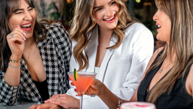 Three women sit at a table indoors, smiling and talking, each holding a pink cocktail in a martini glass.