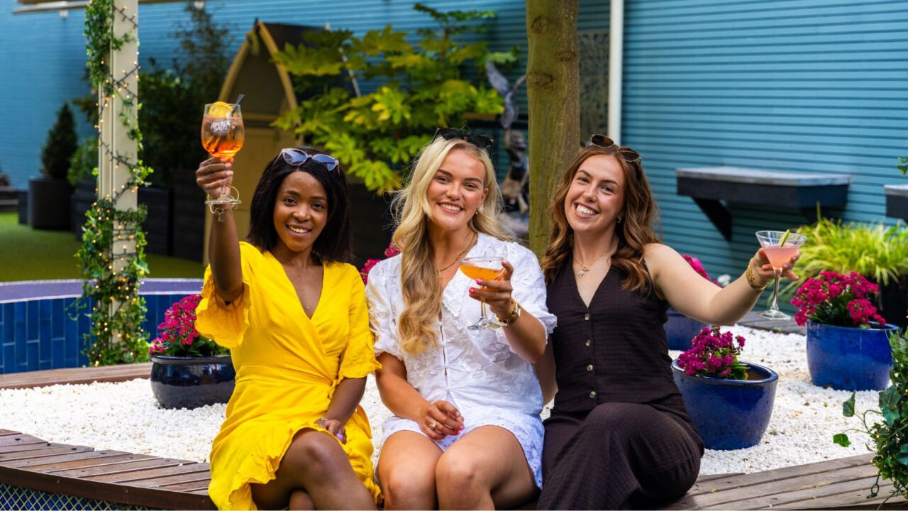 Three women sit outdoors on a wooden bench, smiling and holding up cocktails, with colorful flowers and greenery in the background.