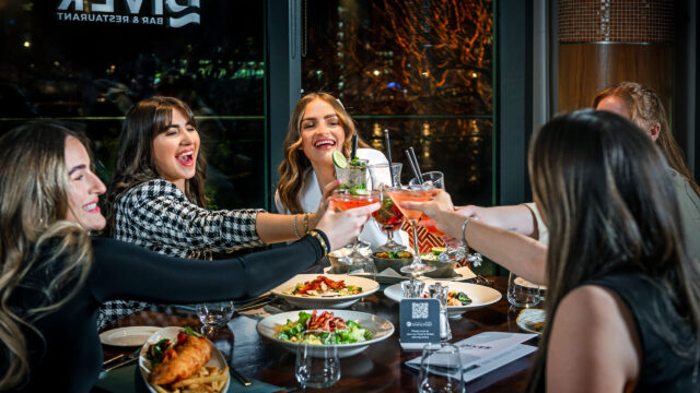 Five women sit around a restaurant table, raising glasses in a toast, with plates of food in front of them and a window showing the restaurant's name in the background.
