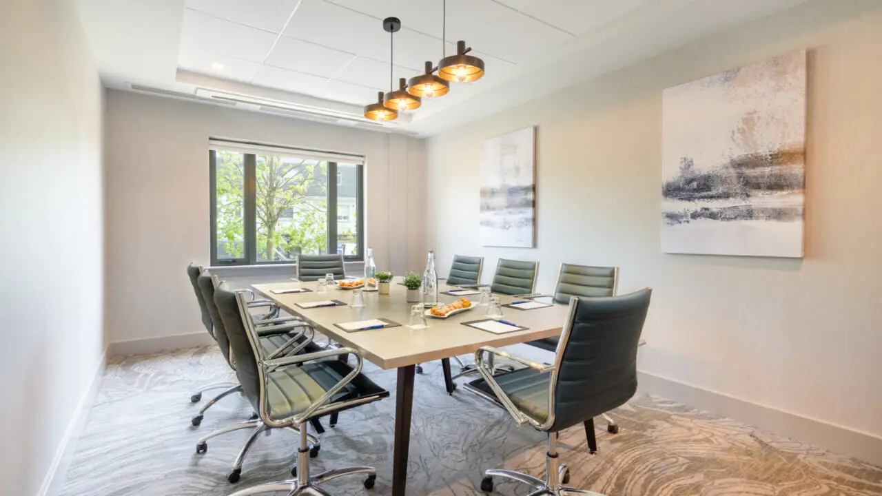Modern conference room with a rectangular table, eight chairs, notepads, water bottles, snacks, and abstract art on the walls, lit by a ceiling fixture and natural light from a window.
