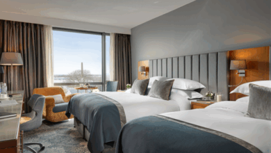 Modern hotel room with two double beds, bedside tables, a desk with a chair, a lounge chair by the window, and a view of trees and buildings outside.