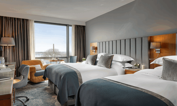 Modern hotel room with two double beds, bedside tables, a desk with a chair, a lounge chair by the window, and a view of trees and buildings outside.