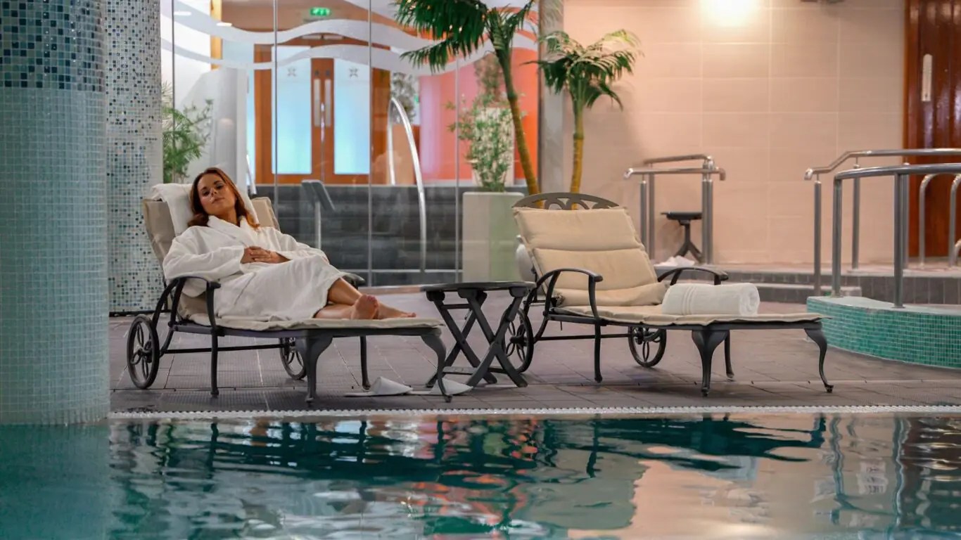 A woman in a white robe relaxes on a lounge chair beside an indoor pool, with towels and a table nearby.