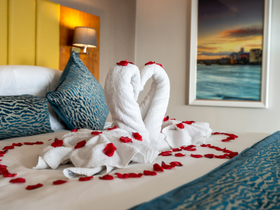 Two towel swans forming a heart are arranged on a bed with blue pillows, surrounded by red rose petals, in a modern hotel room.