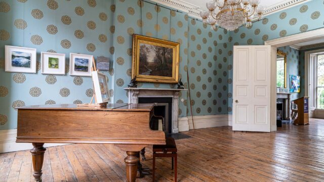 A grand piano sits on a wooden floor in an elegant room with blue patterned wallpaper, a fireplace, framed art, a chandelier, and an open door leading to another space.