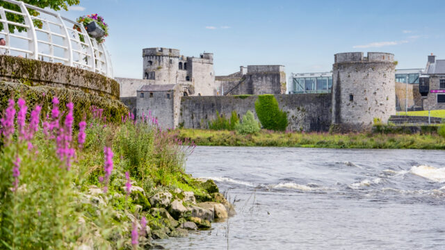 View of a stone castle with round towers beside a river, surrounded by purple wildflowers and greenery under a blue sky&mdash;perfect scenery for memorable Spring Hotel Breaks.