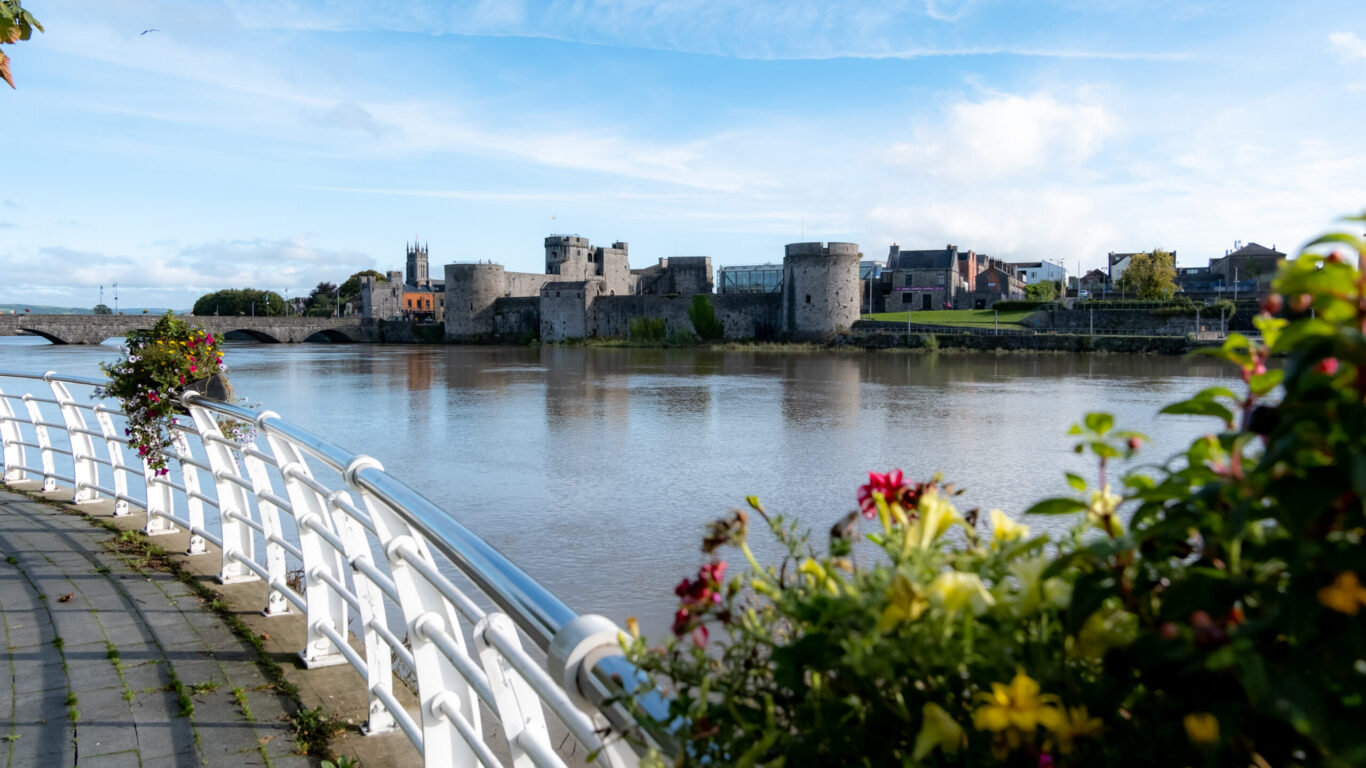 View of a stone castle and buildings across a river, with a curved railing and colorful flowers in the foreground under a partly cloudy sky&mdash;a perfect scene for relaxing spring hotel breaks.