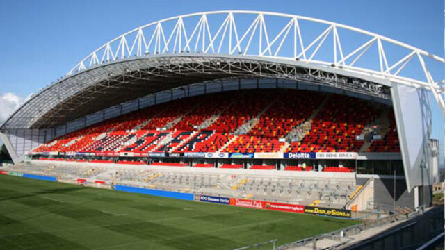 A modern rugby stadium with red, orange, and black seating under a large arched roof, adjacent to a well-maintained grass field.