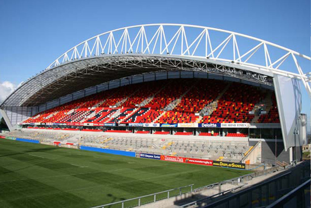 A modern rugby stadium with red, orange, and black seating under a large arched roof, adjacent to a well-maintained grass field.