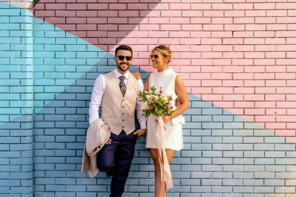 A man in a beige vest and dark pants stands beside a woman in a white dress holding a bouquet; both wear sunglasses and stand against a colorful, geometric brick wall. The best hotel wedding packages in Limerick at the Limerick Strand Hotel.