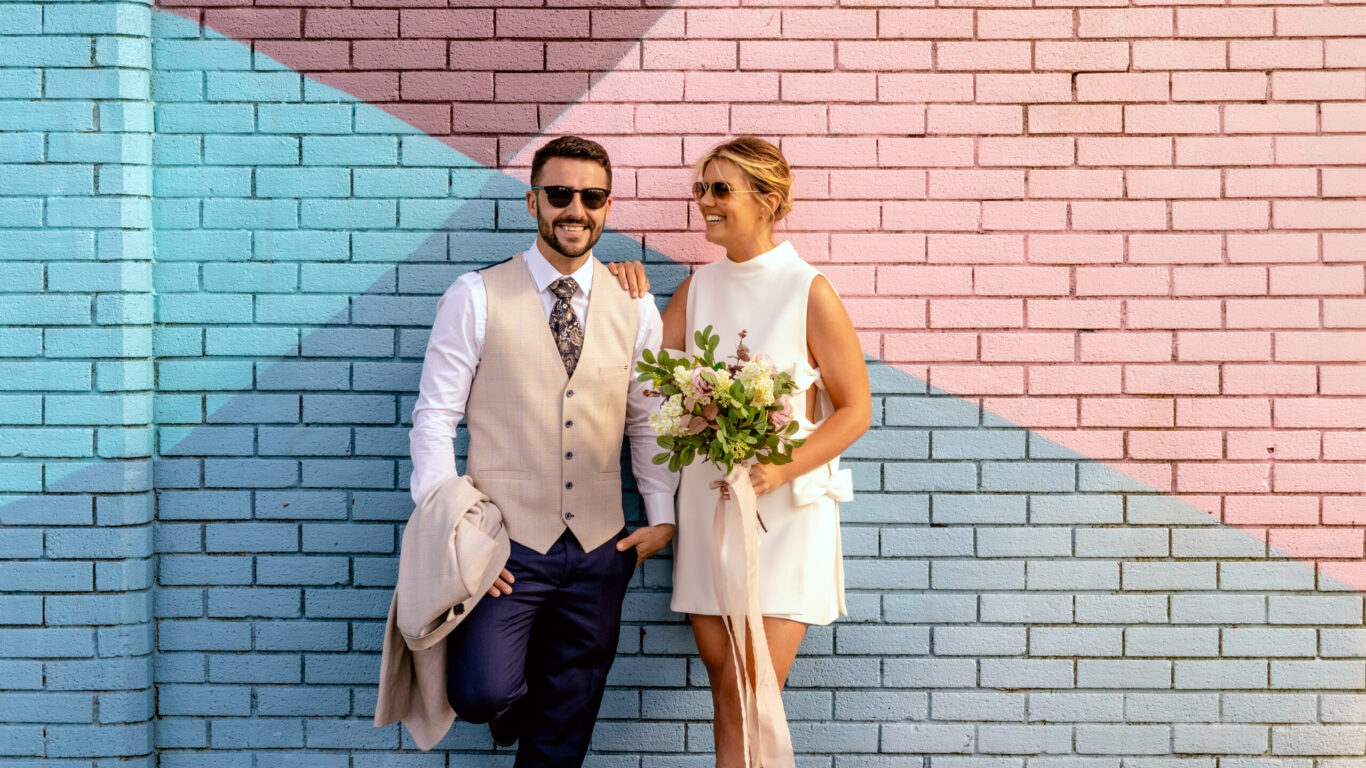A man in a beige vest and dark pants stands beside a woman in a white dress holding a bouquet; both wear sunglasses and stand against a colorful, geometric brick wall. The best hotel wedding packages in Limerick at the Limerick Strand Hotel.