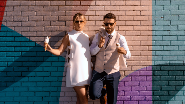 A man and woman dressed formally lean against a colorful brick wall, both wearing sunglasses and holding ice cream cones.