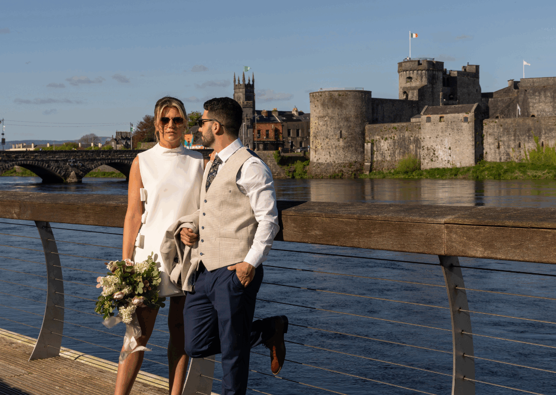 A couple dressed in formal attire stands by a riverside boardwalk, with a bouquet and historic stone castle in the background under a clear blue sky.