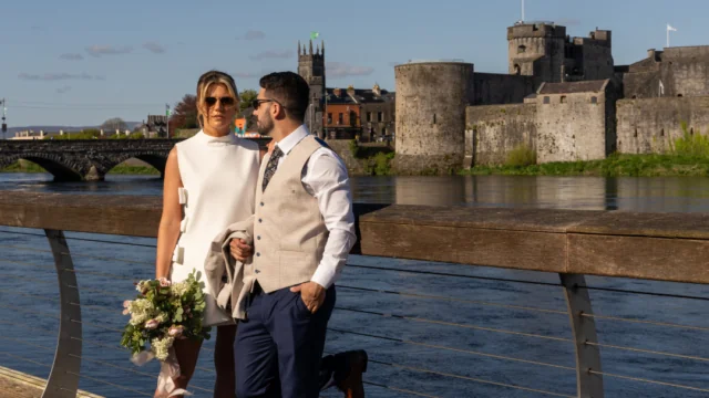 A couple dressed in formal attire stands by a riverside boardwalk, with a bouquet and historic stone castle in the background under a clear blue sky.