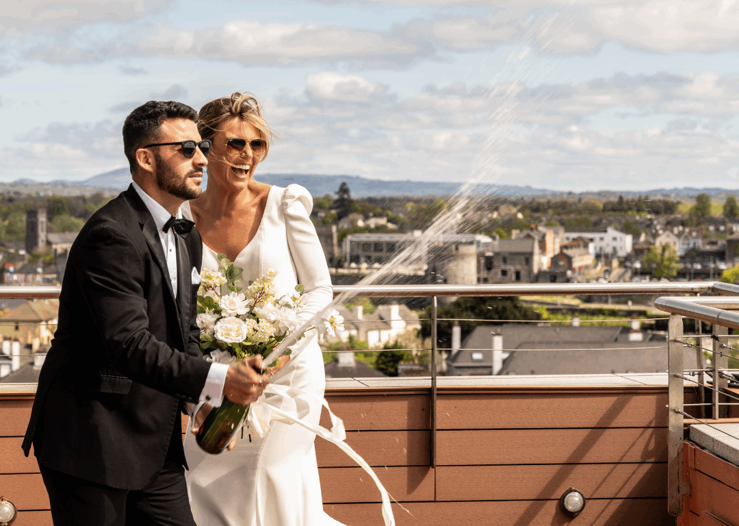 A bride and groom in formal attire stand on a rooftop, smiling as the groom pops a bottle of champagne, with city buildings and a cloudy sky in the background.